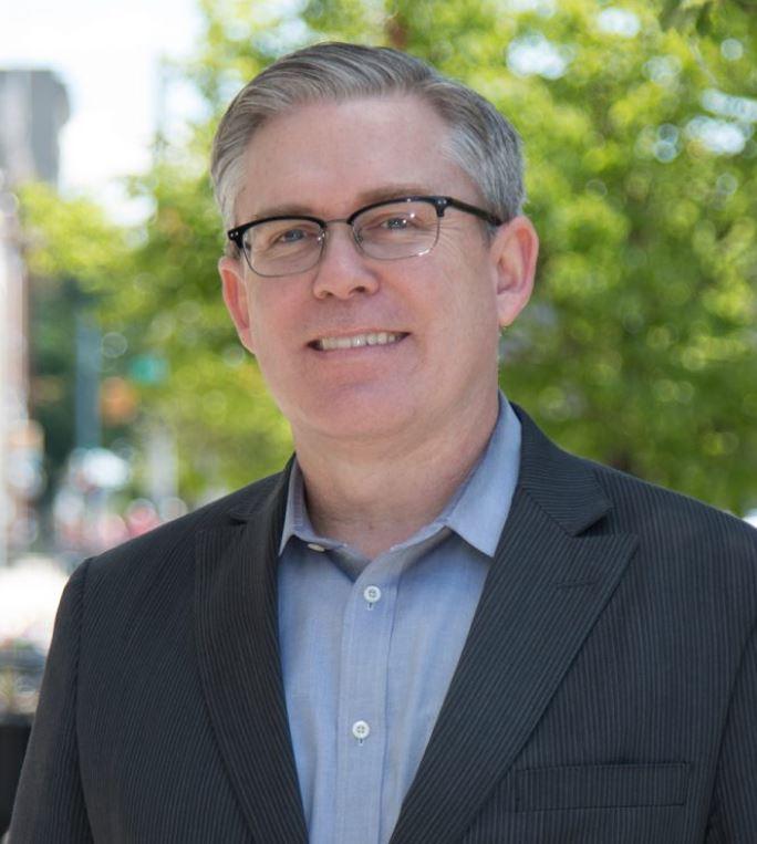 Portrait of Neal Hegarty in a light blue shirt and dark blue Jacket, wearing glasses.