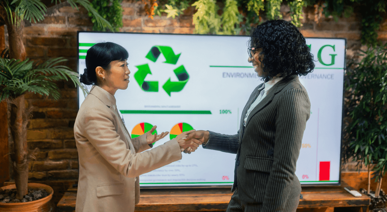 Two women of color in suits shaking hands infront of a presentation screen.