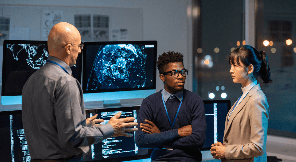 Three people in an office talking in front of computer screens.
