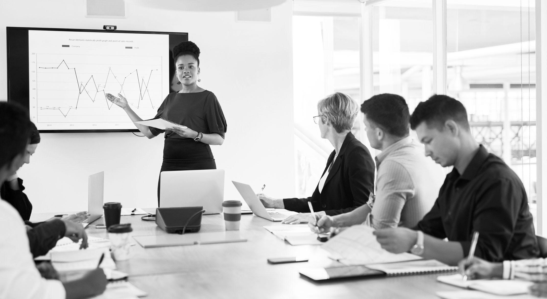 a group of businesspeople in a meeting in a modern office; a woman of color presenting in front of a screen with data charts.
